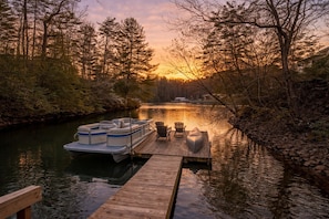 A wooden dock extending into a calm river, with a boat docked at the end and a row of chairs and a canoe nearby.