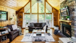 A cozy cabin interior with a wooden ceiling, a fireplace, a large window, and a leather sofa.