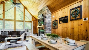 A cozy cabin interior with a stone fireplace, wooden walls, a dining table set for four, and a comfortable seating area with a couch and coffee table.