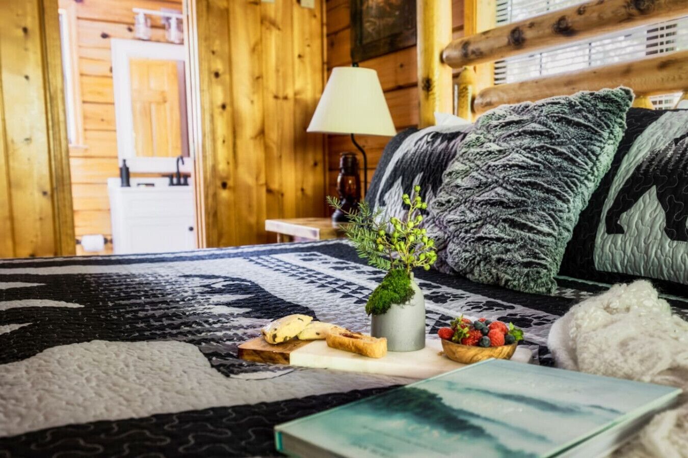 Une pièce chaleureuse, avec des murs en bois, comprenant un lit, une lampe, un vase avec des plantes et un plateau de nourriture.