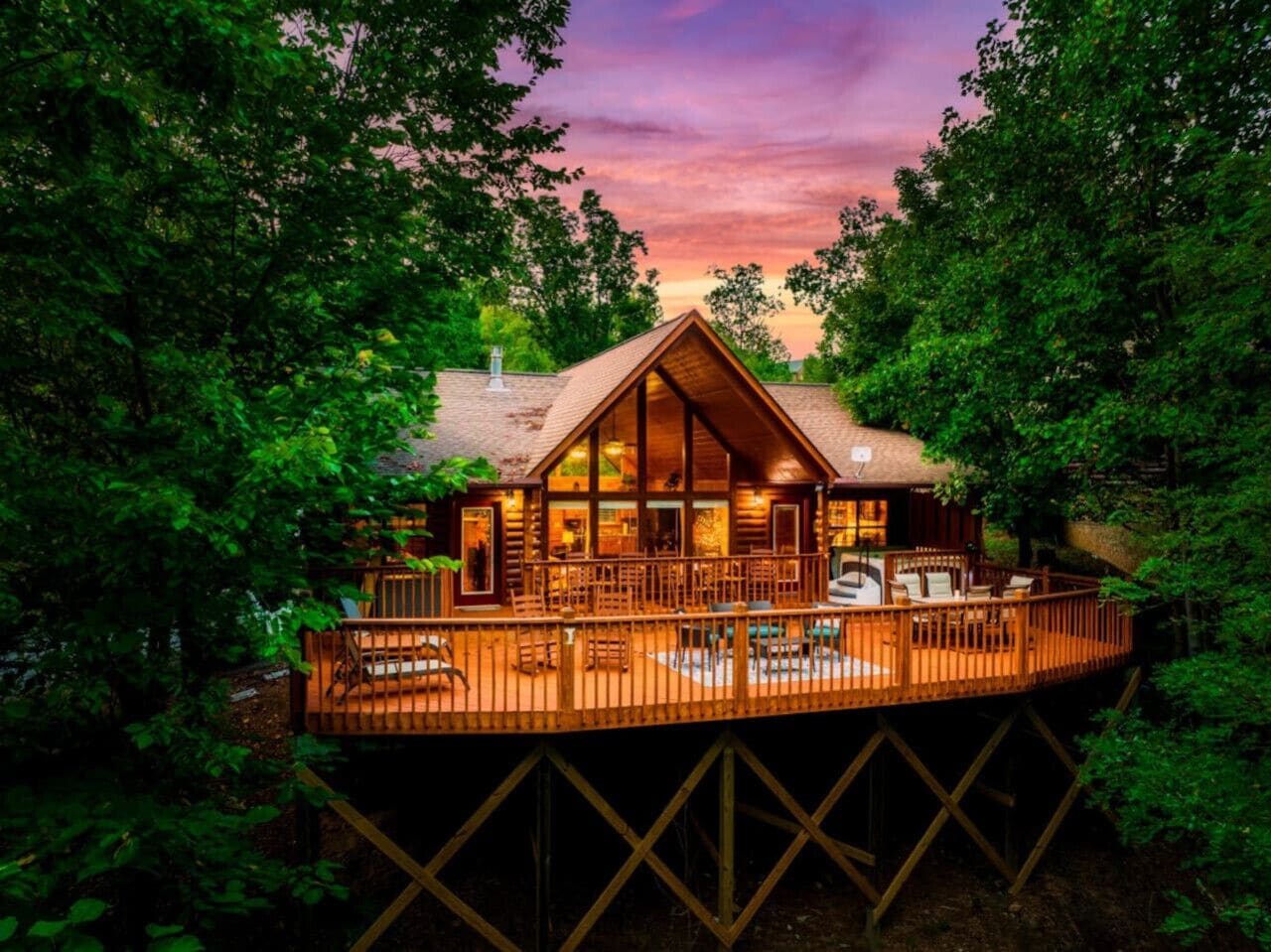 A wooden cabin with a deck, surrounded by trees, at sunset.
