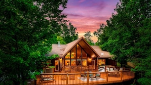 A wooden cabin with a deck, surrounded by trees, at sunset.