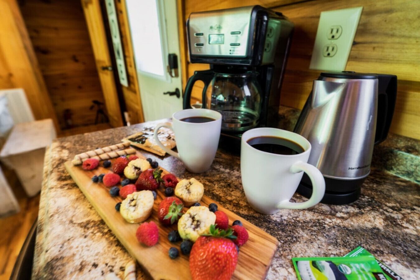 A breakfast setup with a coffee maker, two cups of coffee, a cutting board with pastries and fresh fruits, and a kettle.