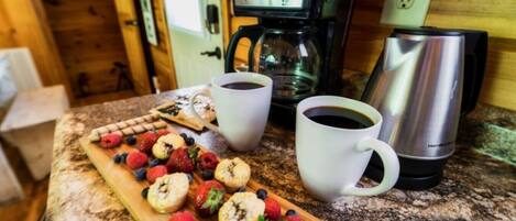A breakfast setup with a coffee maker, two cups of coffee, a cutting board with pastries and fresh fruits, and a kettle.