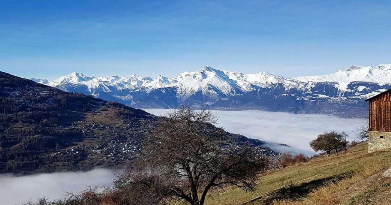 A mountainous landscape with snow-capped peaks, a valley with a small settlement, and a wooden building on the right.