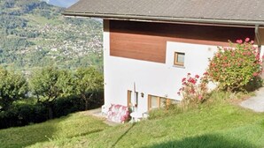 A two-story house with a sloped roof, a chimney, and a view of a valley with mountains in the background. - Petit Appartement à Louer Dans le Val Dhérens (Vernamiège)