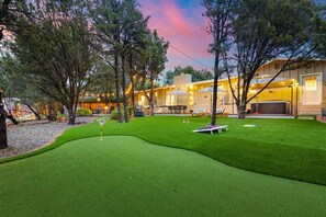Um gramado bem cuidado com putting green, área de estar externa e um edifício iluminado com luzes quentes.