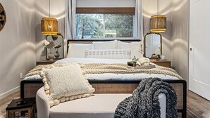 Un dormitorio con una cama grande, dos lámparas de noche, una ventana con vistas a árboles y una mesita de noche con un florero y una planta.