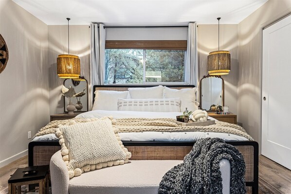 A bedroom with a large bed, two bedside lamps, a window with a view of trees, and a bedside table with a vase and a plant. - Sedona Villa•Magical Backyard •Fire Pit•Hot Tub•Near Trails (Sedona)