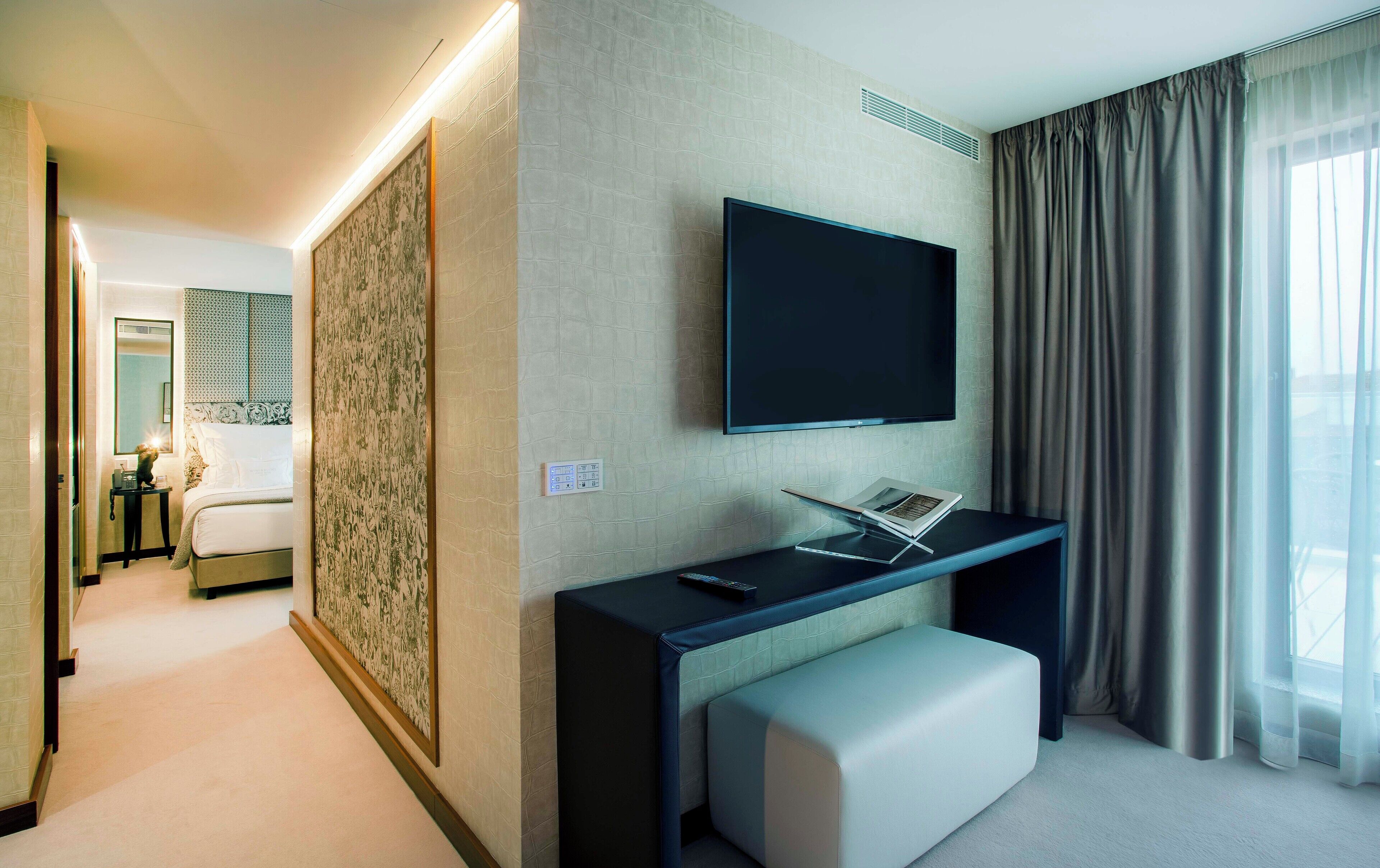 A modern hotel room with a flat-screen TV mounted on the wall, a bedside table with a remote control, a large window with curtains, and a hallway leading to another room.