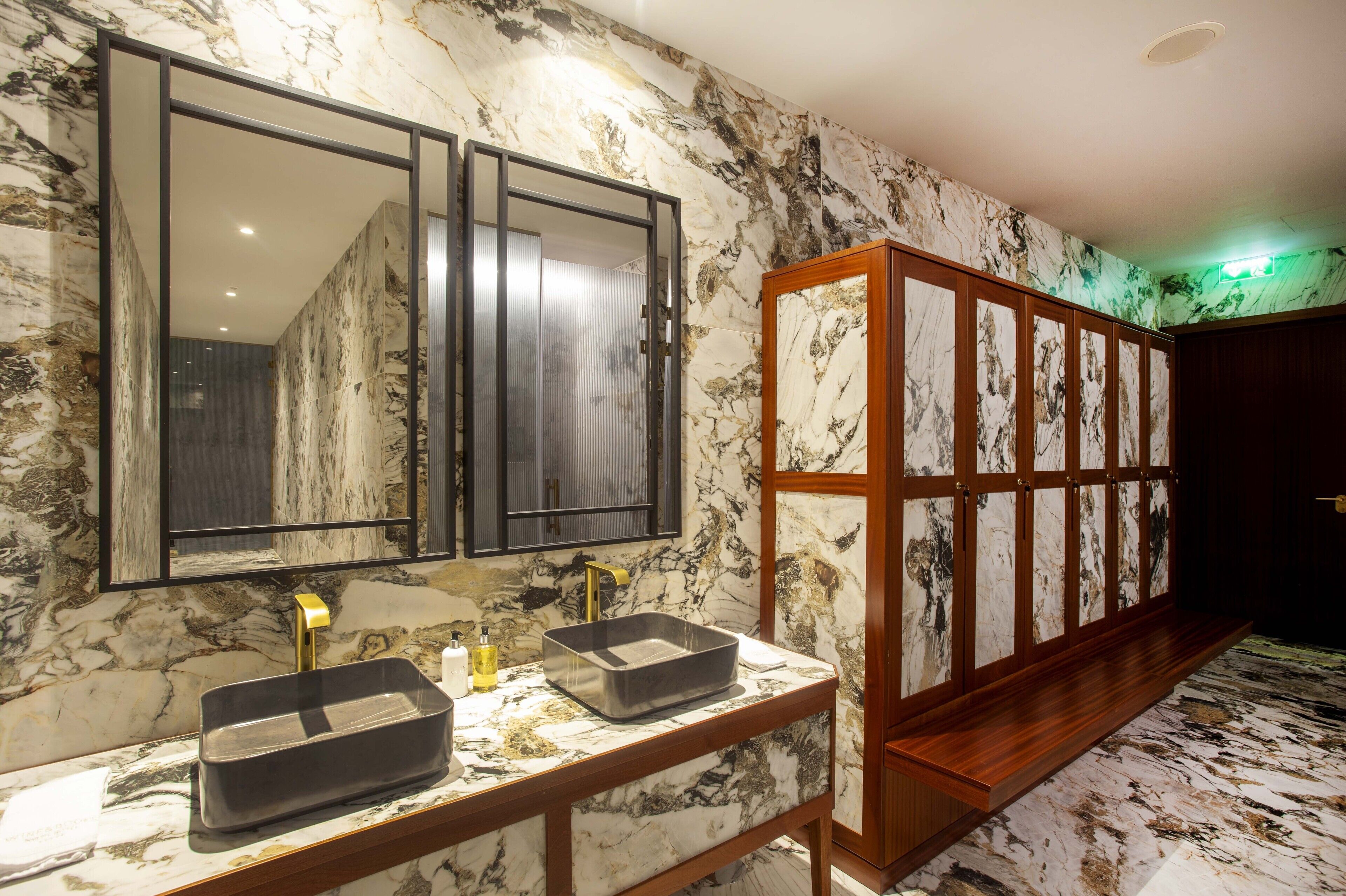 Une salle de bain avec un mur en marbre, un grand miroir, un lavabo avec un robinet doré et un banc en bois.