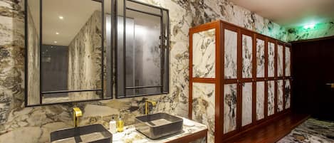 Une salle de bain avec un mur en marbre, un grand miroir, un lavabo avec un robinet doré et un banc en bois.
