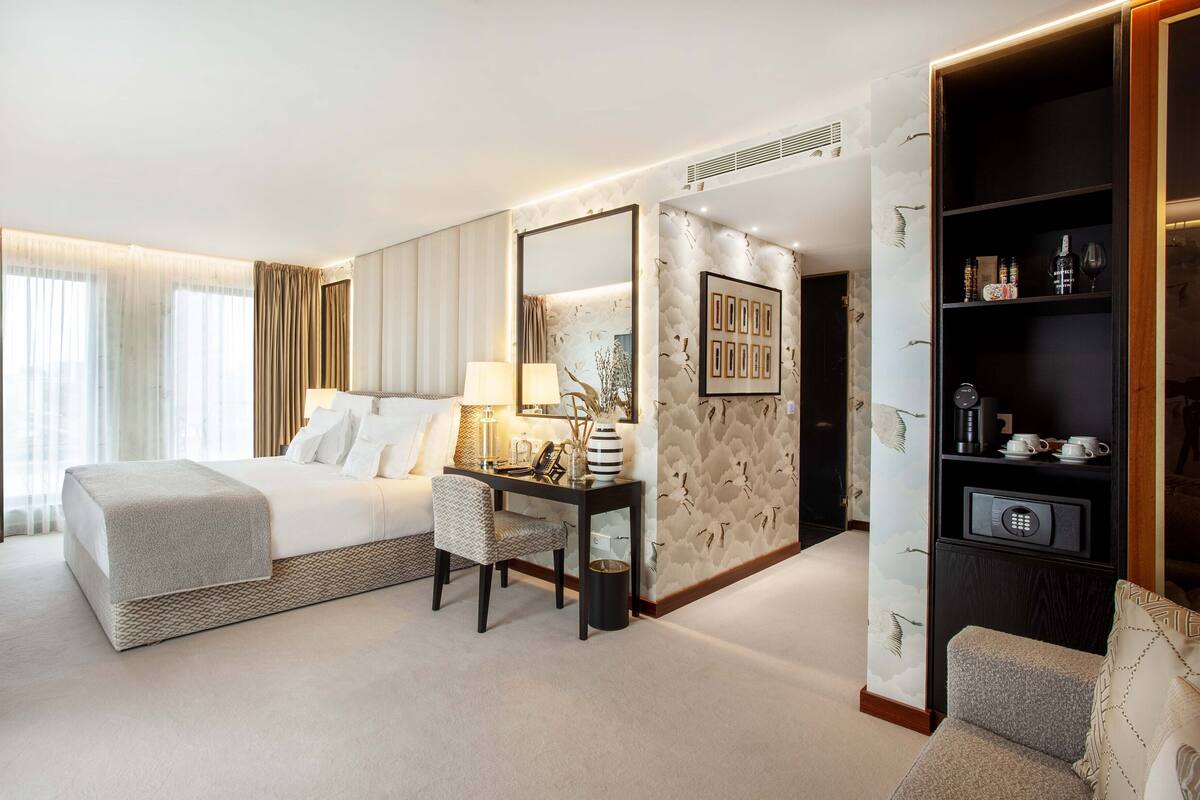 A hotel room with a bed, bedside table, lamp, and a large mirror. There is a wall with a decorative panel and a bookshelf with various items.