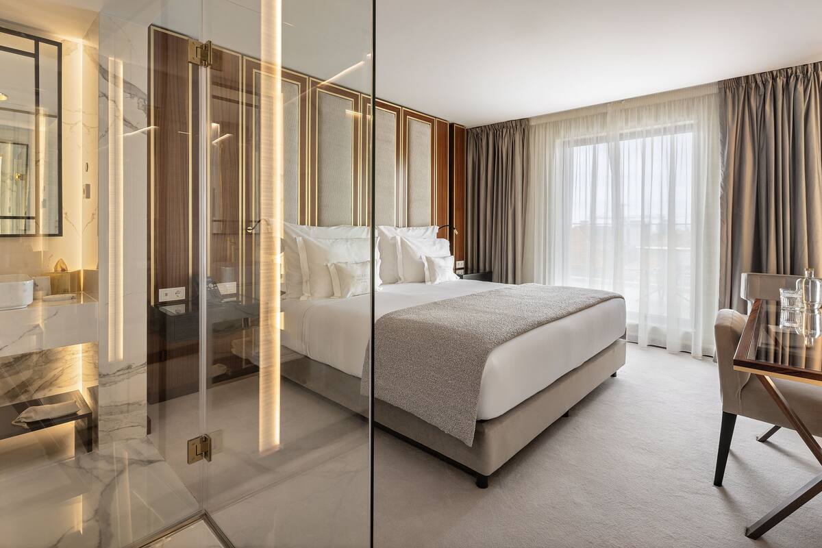 A modern hotel room with a large bed, a glass partition, a desk with a chair, and a view of the outdoors through sheer curtains.