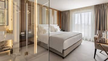 A modern hotel room with a large bed, a glass partition, a desk with a chair, and a view of the outdoors through sheer curtains.