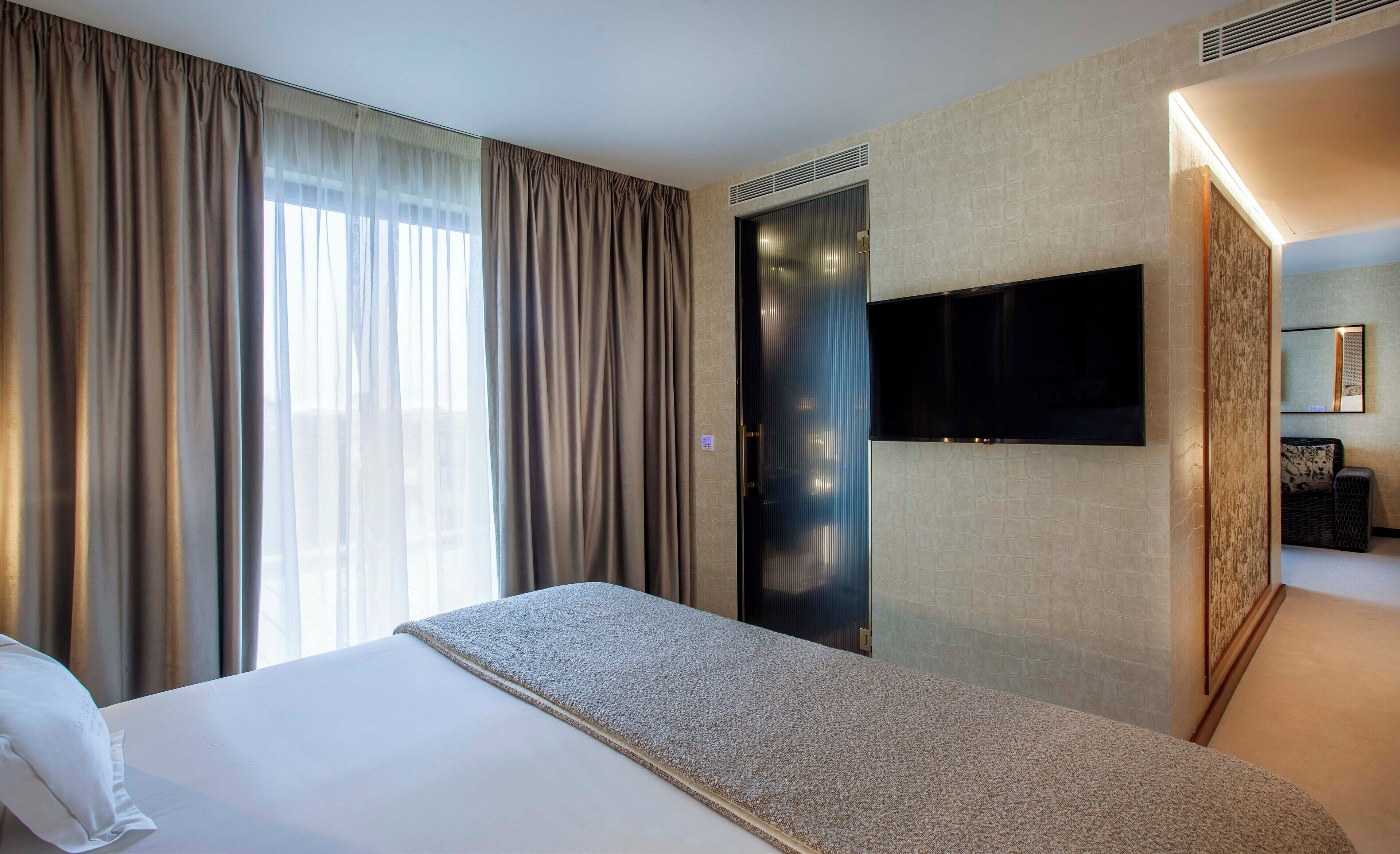 A hotel room with a large bed, a flat-screen TV mounted on the wall, and a large window with curtains.