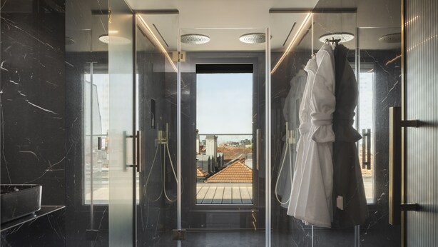 A modern bathroom with a glass shower enclosure, a visible cityscape through the door, and a hanging robe. - Wine & Books Porto Hotel - Small Luxury Hotels of the World (Porto)