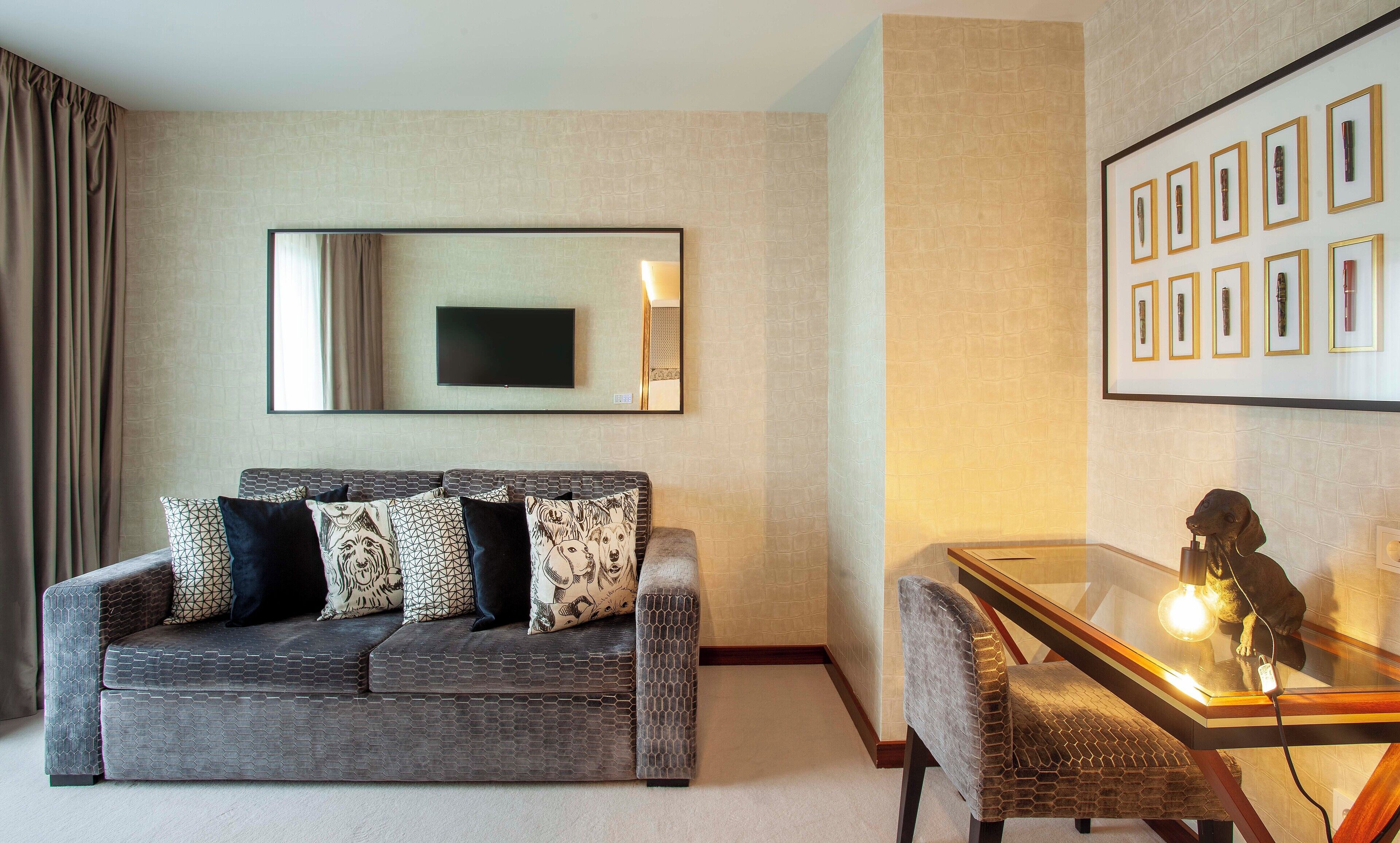 A modern living room with a grey sofa, patterned cushions, a flat-screen TV, and a glass coffee table with a sculpture.