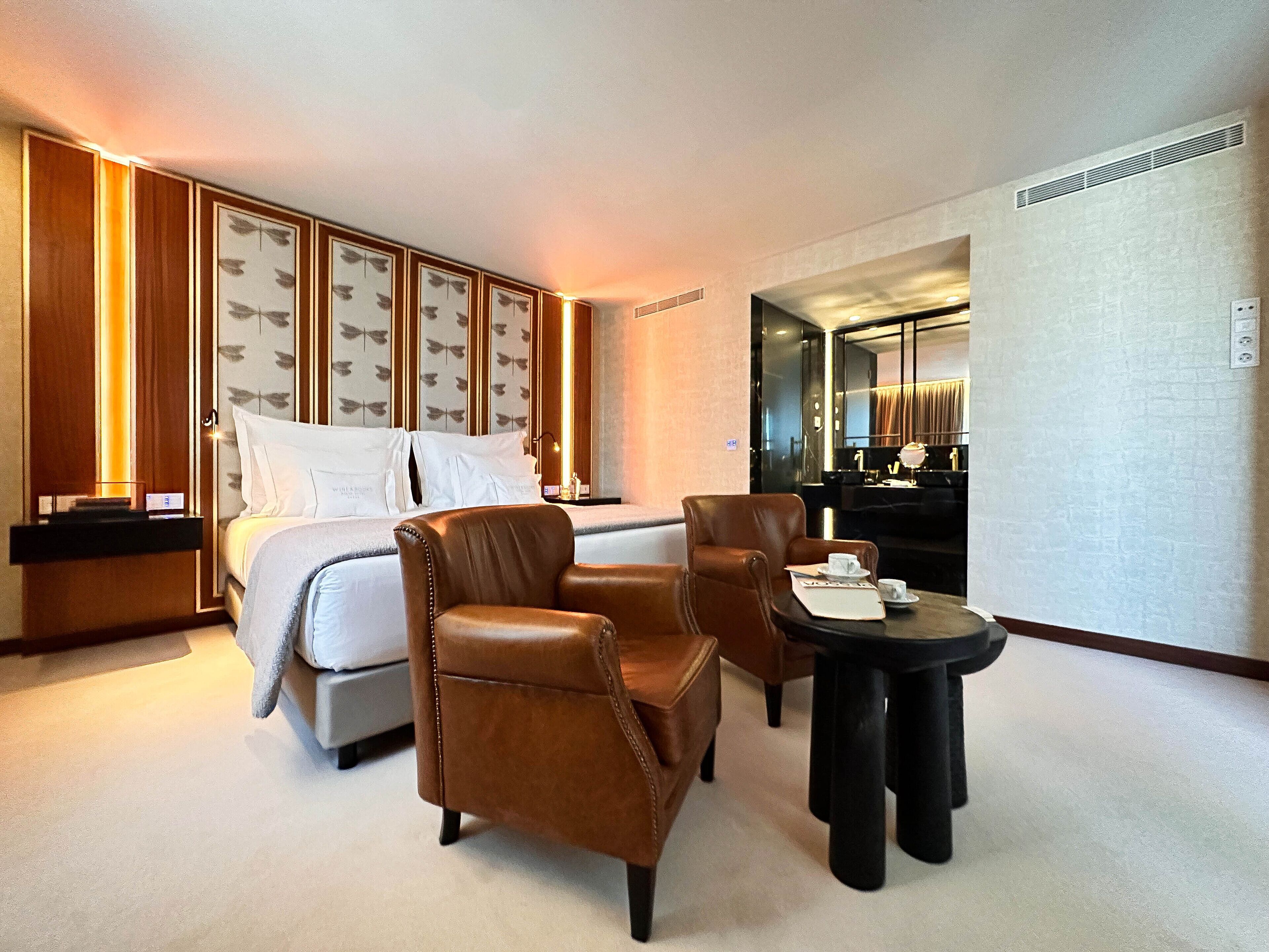 A hotel room with a bed, two leather armchairs, a small table with a cup and saucer, and a mirror.