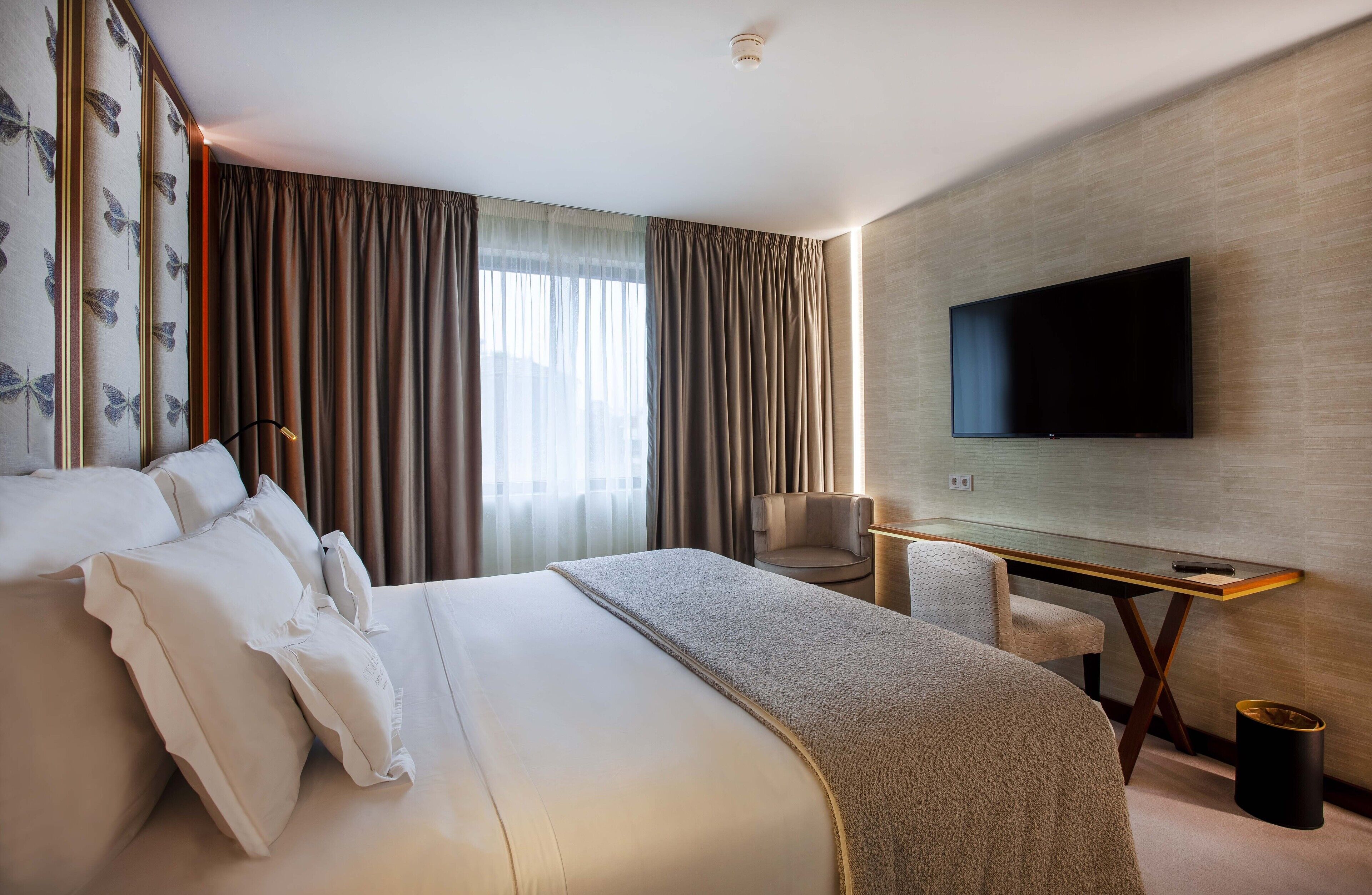 A modern hotel room with a large bed, a desk, a chair, a flat-screen TV, and a large window with curtains.