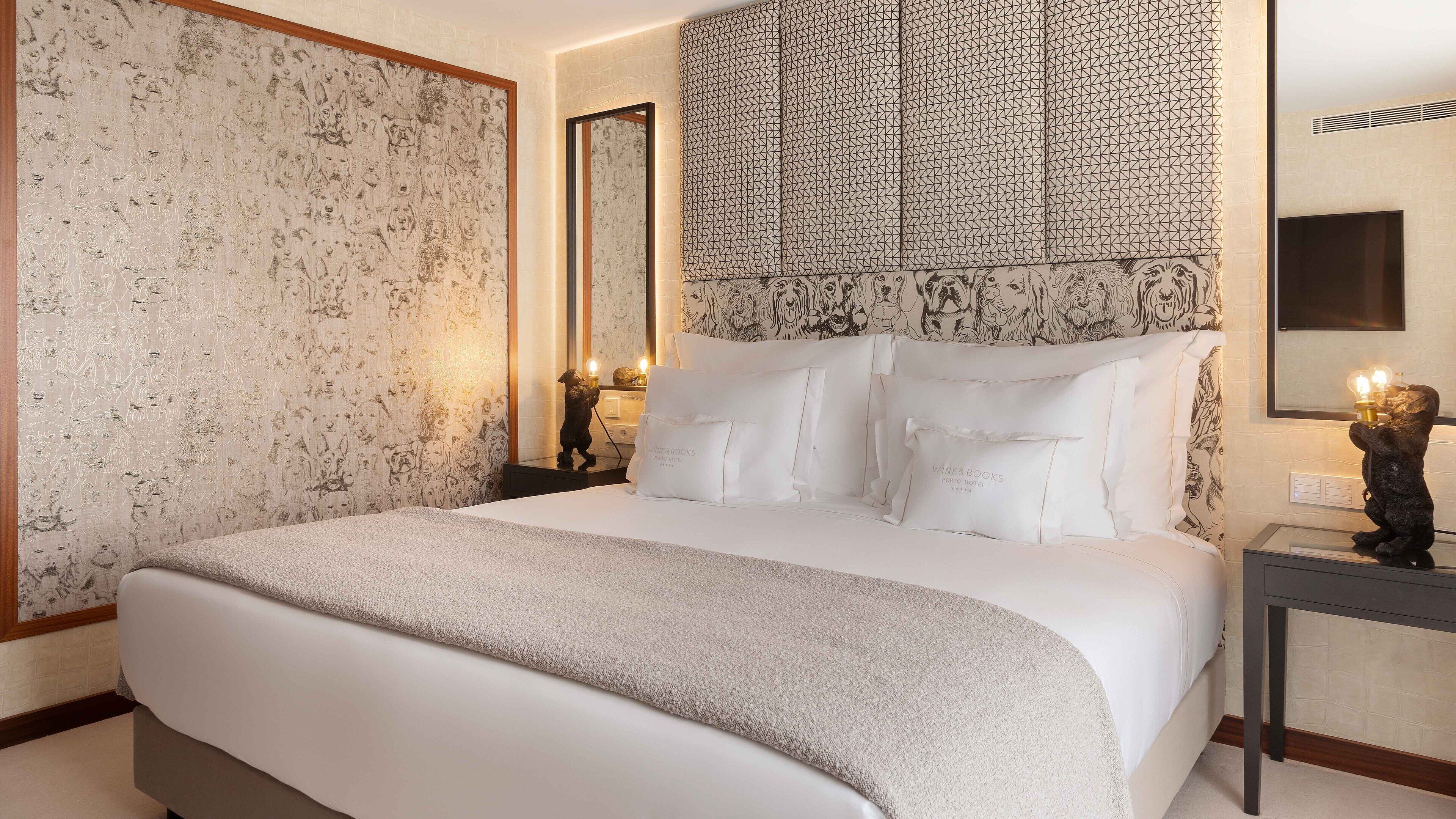 A neatly made bed with white linens and pillows, a bedside table with a lamp, and a wall with a geometric patterned wallpaper.