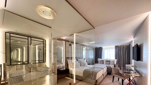 A modern hotel room with a large mirror, a separate bathroom, a bed with multiple pillows, a small table, and a TV mounted on the wall. - Wine & Books Porto Hotel - Small Luxury Hotels of the World (Porto)