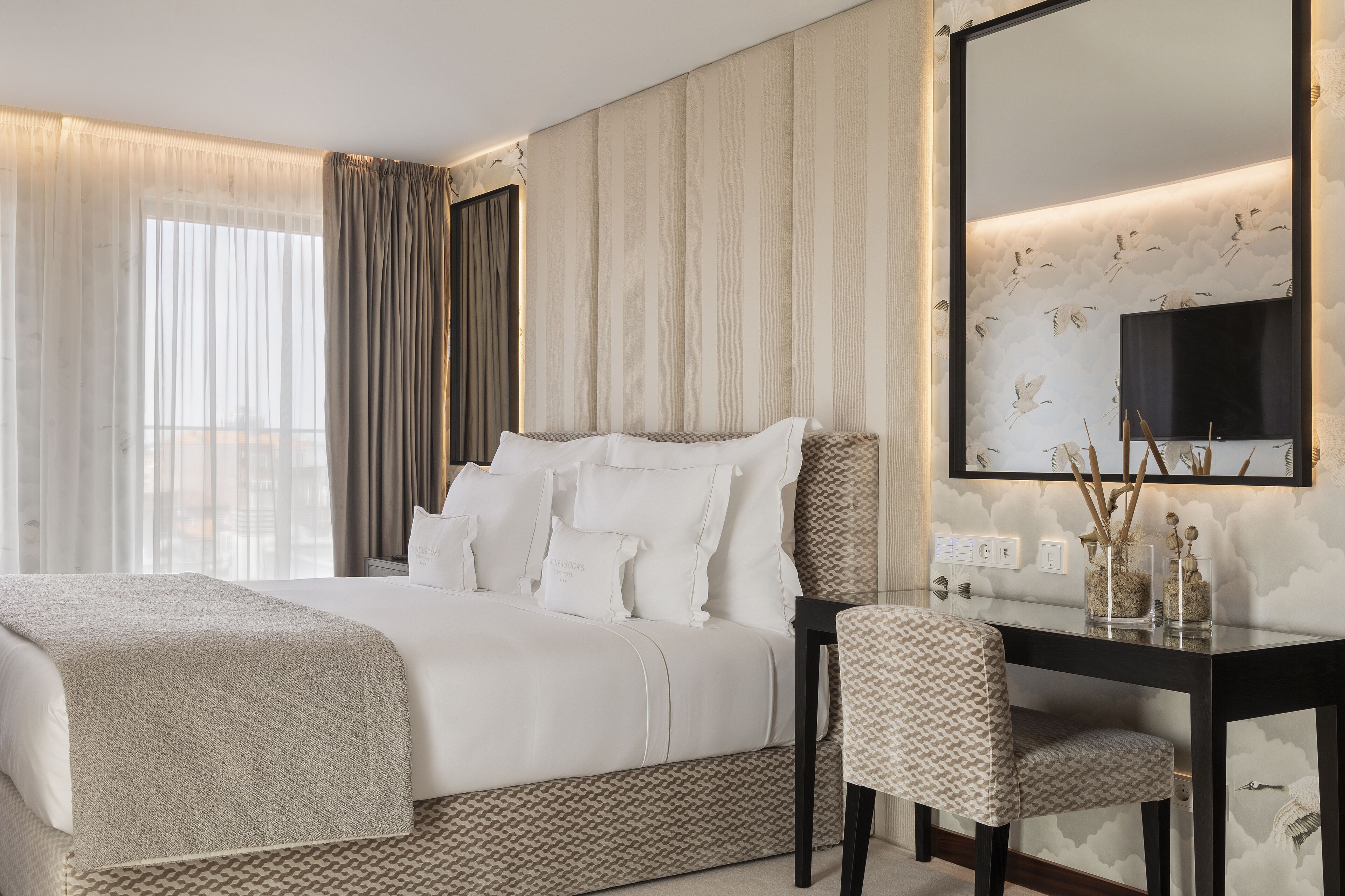 Une chambre d’hôtel avec un grand lit, un bureau et une chaise. Il y a un grand miroir au mur et une fenêtre avec des rideaux.