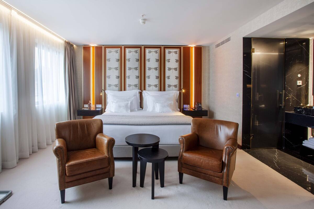 A modern hotel room with a bed, two leather armchairs, a small round table, and a minibar.