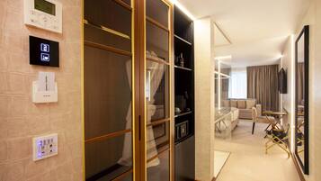 Quarto de hotel moderno com porta de correr de vidro, roupeiro embutido, área de estar com sofá e poltronas, e um espelho grande na parede.