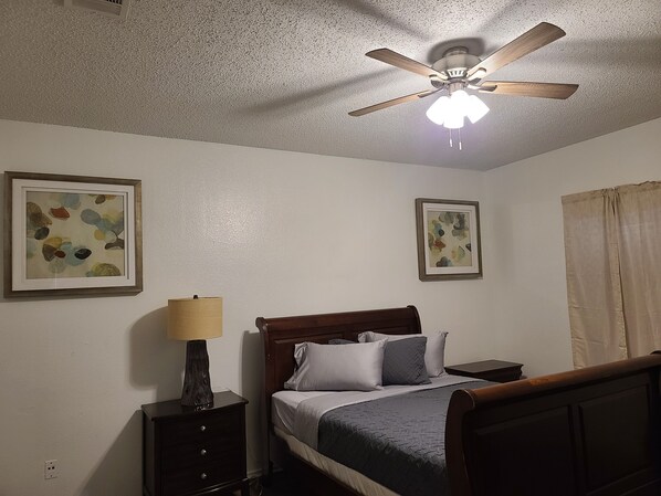 Ein Schlafzimmer mit einem Bett, einem Nachttisch, einer Lampe, zwei gerahmten abstrakten Bildern und einem Deckenventilator.