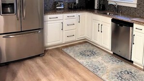 Una cocina con armarios blancos, un refrigerador de acero inoxidable, un lavavajillas y un ventanal con vista a árboles.