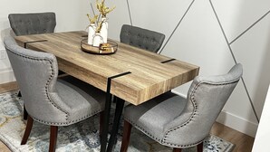 Comedor con mesa de madera, cuatro sillas grises tapizadas, alfombra estampada y un reloj de pared.