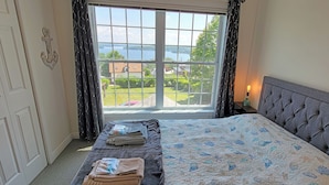 Een slaapkamer met een groot raam, een bed met een grijs hoofdeinde, en een nachtkastje met een lamp.