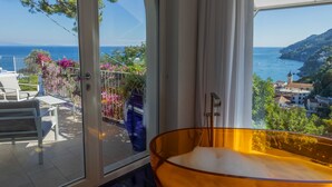 Habitación con un gran ventanal que ofrece vistas a la costa, una bañera y un balcón con mobiliario exterior y flores.