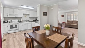 Cozinha moderna com armários brancos, área de jantar com mesa e cadeiras de madeira e sala de estar com sofá bege.