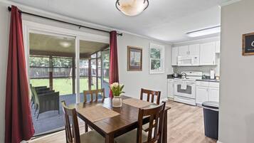 Een eetgedeelte met een houten tafel en stoelen, een vaas met bloemen, en uitzicht op de achtertuin door glazen deuren.