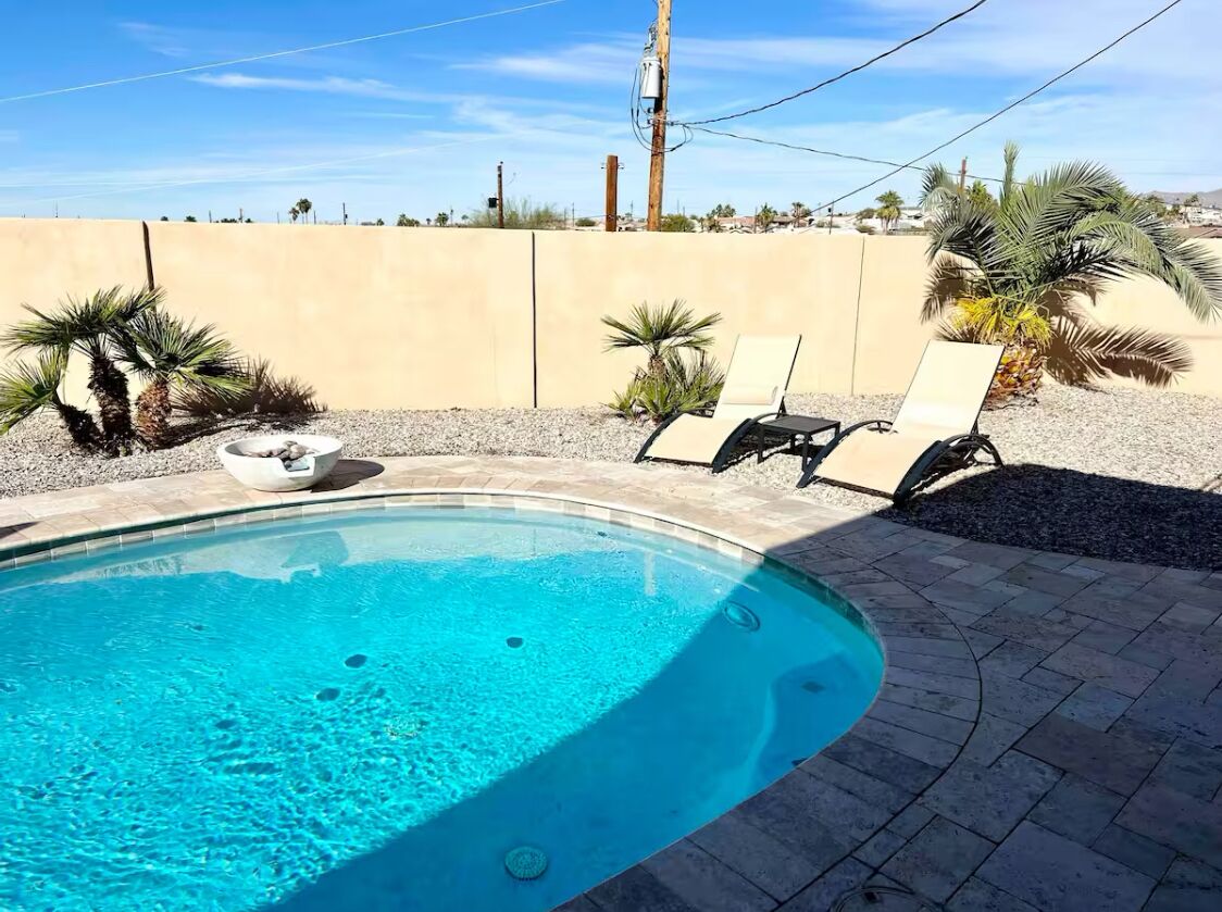 Una piscina con dos sillas reclinables y una mesita, rodeada de palmeras y un muro color beige.