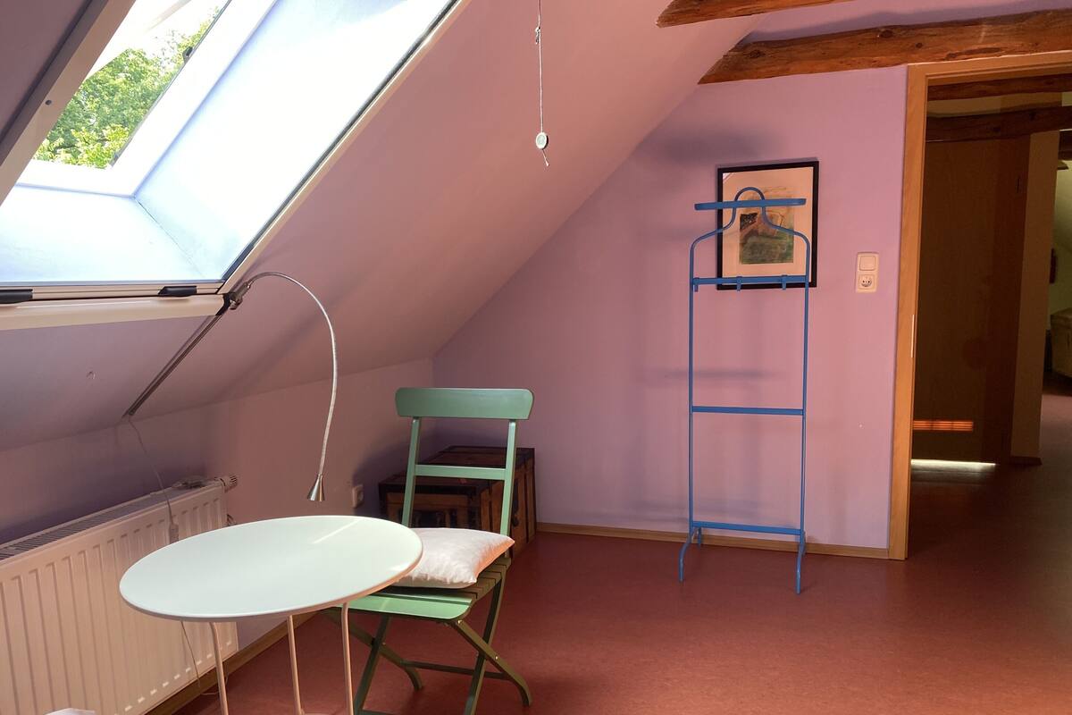 Una habitación con techo inclinado, una mesa con sillas, un radiador y una puerta que da a otra habitación.