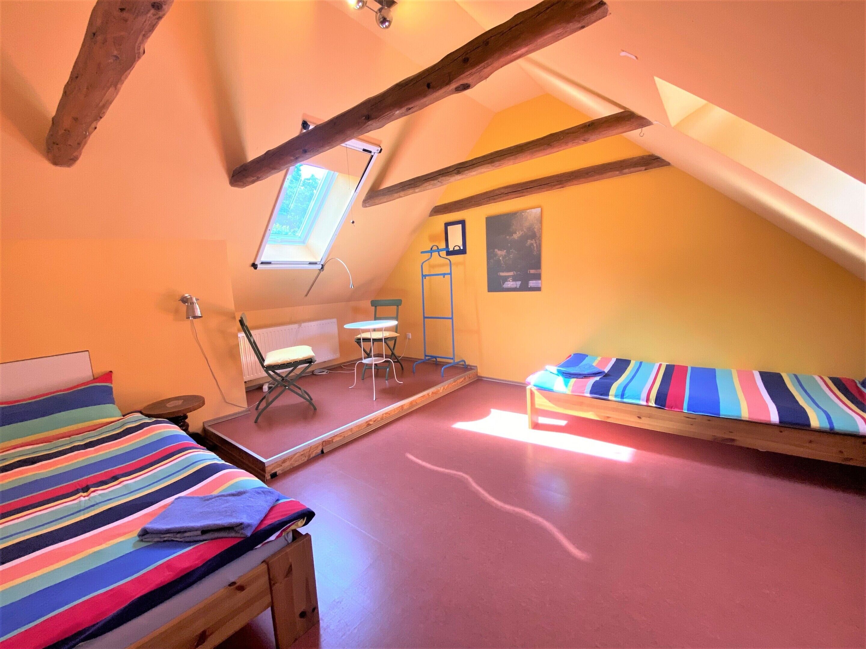 Una stanza piccola e accogliente con soffitto spiovente, una finestra a lucernario, un letto con copriletto a righe colorate, un tavolino con una sedia e un letto a castello con materasso a righe.