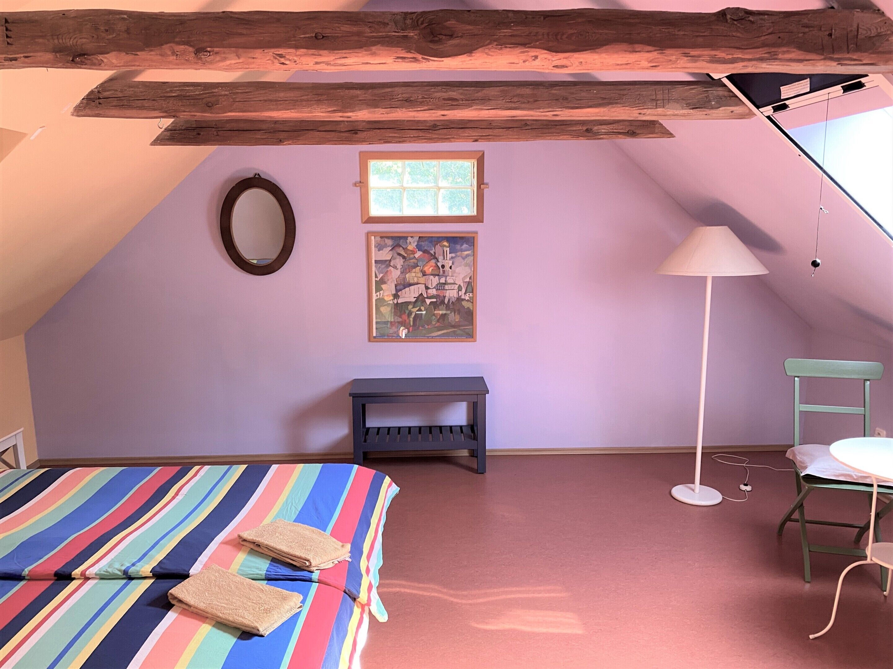 A bedroom with a sloped ceiling, a bed with a striped bedspread, a small bench, a chair, a floor lamp, and a painting on the wall.