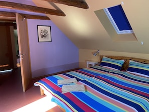 Un dormitorio con techo inclinado, una cama con colcha a rayas coloridas, dos almohadas, una mesita de noche con lámpara y un cuadro en la pared.