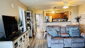 Un salon moderne avec un canapé d’angle gris, un téléviseur à écran plat, des meubles en bois et une cuisine en arrière-plan.
