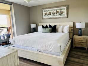 Ein ordentlich bezogenes Bett mit Holzrahmen, zwei Nachttische mit Lampen, ein gerahmtes Kunstwerk über dem Bett und ein Balkon, der durch eine offene Tür sichtbar ist.