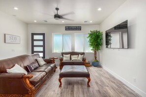 A spacious living room with a brown leather sofa, a matching armchair, and a wooden coffee table. There is a large potted plant, a ceiling fan, and a wall-mounted TV.