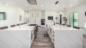 Uma cozinha moderna com ilha central, armários brancos e uma área de jantar com mesa comprida.