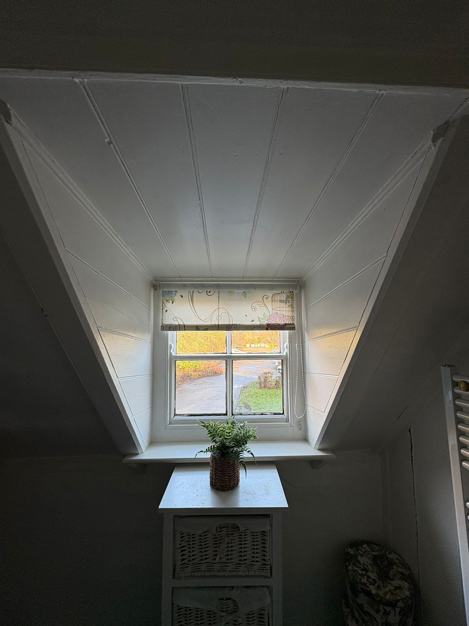 A small attic window with a view of a garden, a potted plant on the sill, and a decorative window valance.