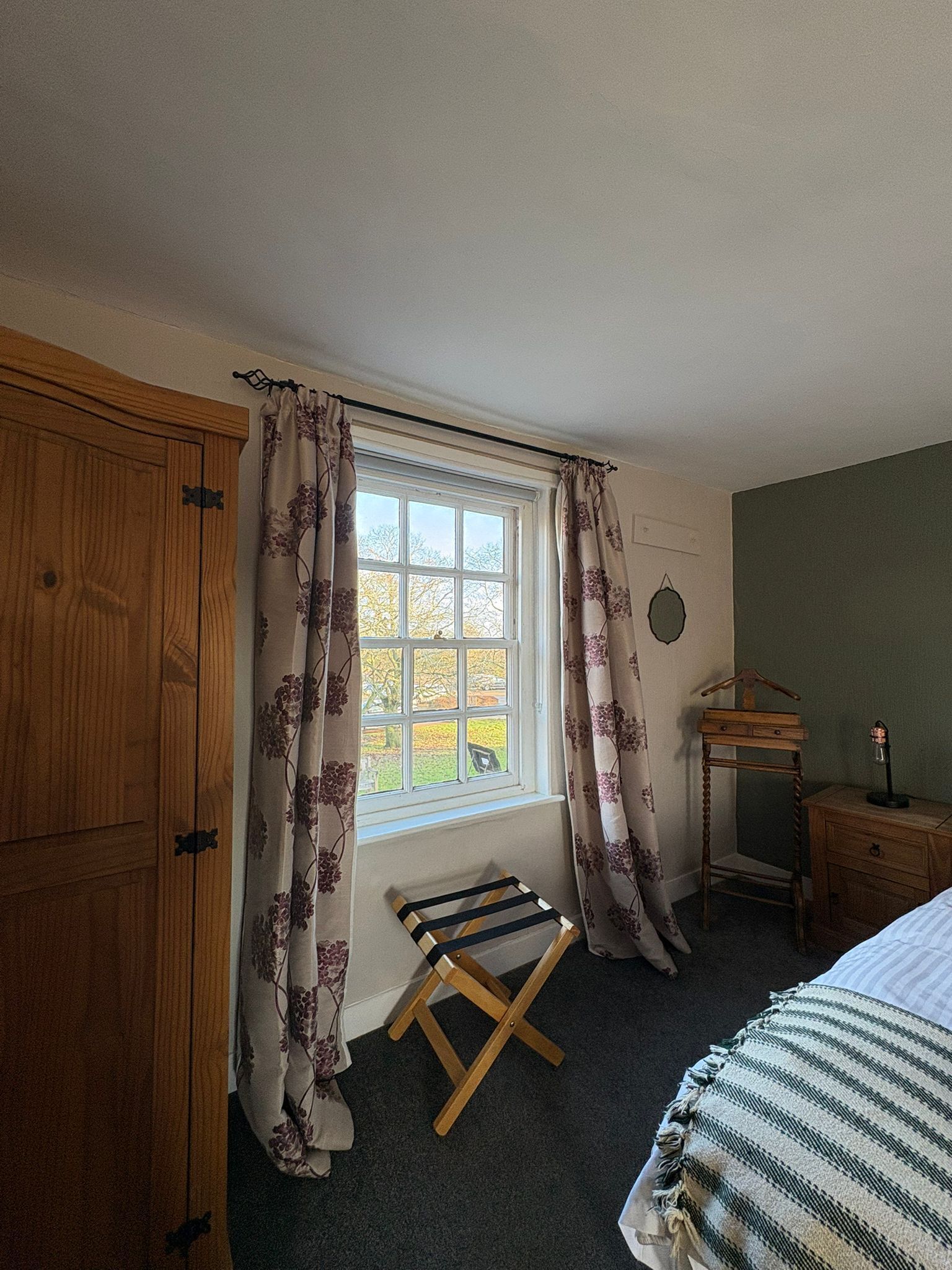A bedroom with a wooden wardrobe, a window with floral curtains, a wooden stool, a bedside table with a lamp, and a striped blanket on the bed.