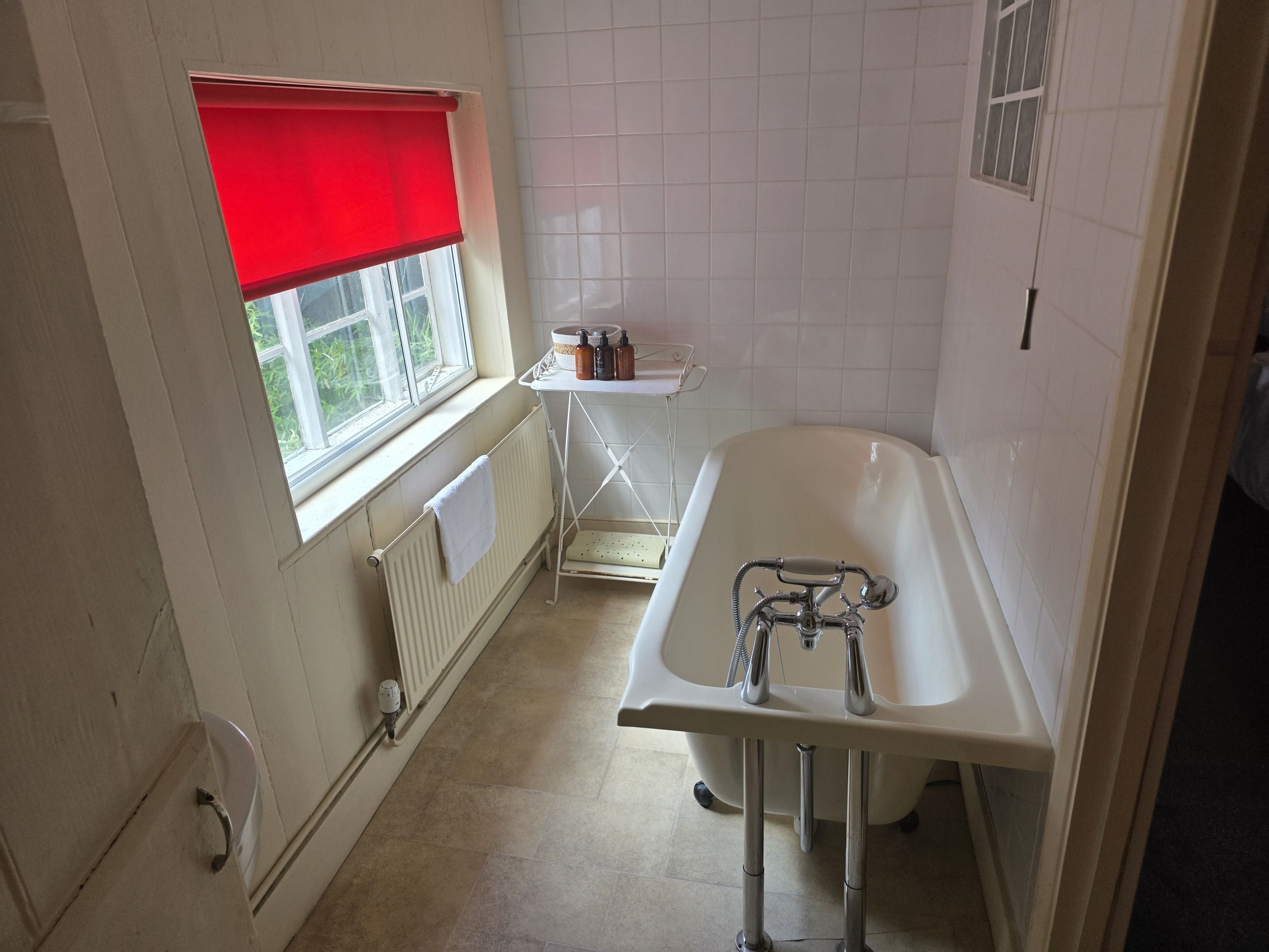 A bathroom with a bathtub, a window with a red blind, a radiator, and a shelf with toiletries.