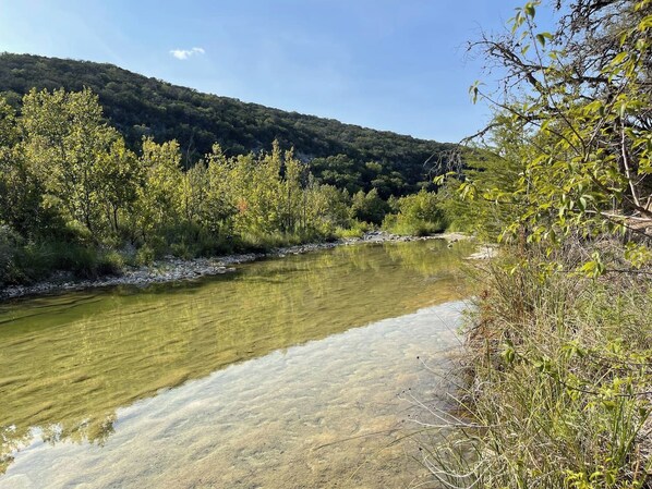 A river with clear water, surrounded by trees and a hill in the background.