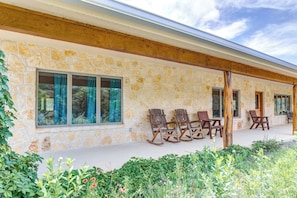 A stone building with a porch, wooden rocking chairs, and a view of the surrounding greenery. - Roadrunner | Riverfront 3BR with Views & Fire Pit (Leakey)
